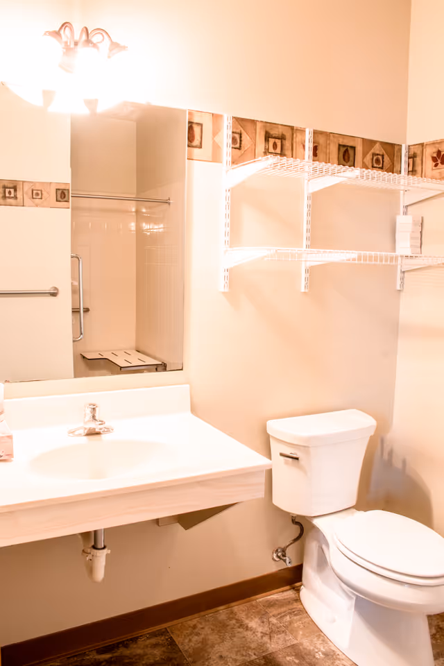 A bathroom with a white sink and faucet, a large mirror above the sink, a white toilet, and white wire shelving mounted on the wall. The walls are beige with a decorative tile border near the ceiling. The floor has brown tiles, and a shower area with a grab bar and a shower bench is visible in the mirror reflection.
