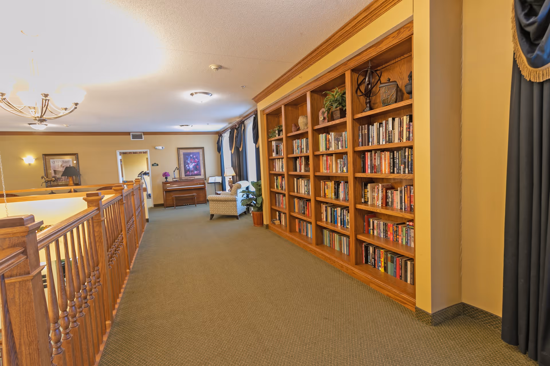 Interior hallway of a senior living facility with a large wooden bookshelf filled with books on the right side, a green carpeted floor, wooden railing on the left, and a seating area with a piano and armchair in the background.