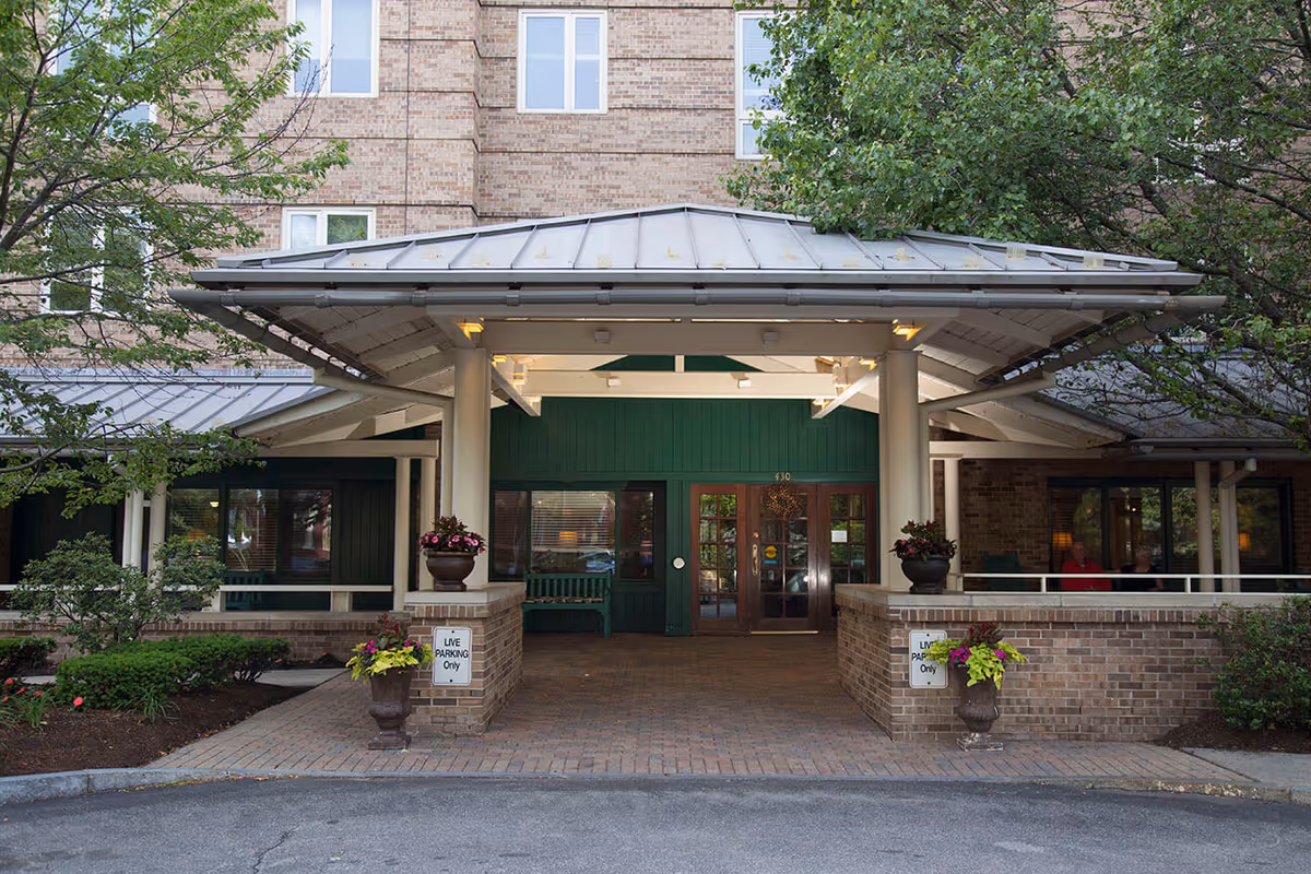 Entrance to a senior living facility with a covered drop-off area supported by white columns. The building is made of brick with green accents around the doors and windows. There are potted plants with flowers on either side of the entrance and small trees and shrubs surrounding the area. Signs indicate 'LIVE PARKING Only'.