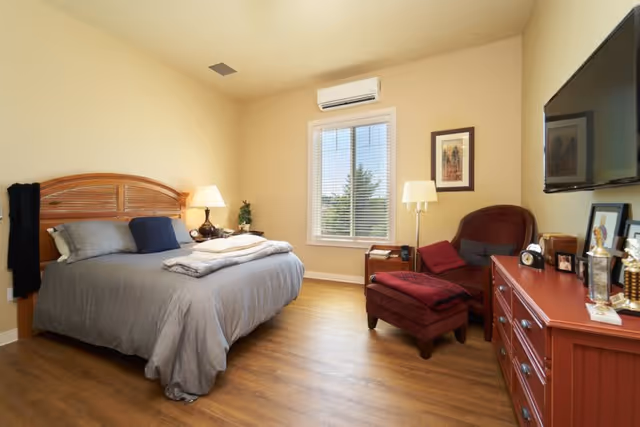A cozy bedroom with a wooden bed frame, gray bedding, and a bedside table with a lamp. There is a window with blinds, a red armchair with a matching ottoman, a floor lamp, a red dresser with various decorative items, and a wall-mounted TV.