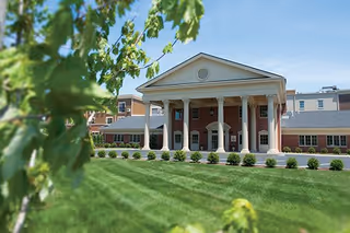 Front exterior view of The Colonnade Senior Living building with a large portico supported by white columns, a well-maintained lawn, and some green leaves in the foreground under a clear blue sky.