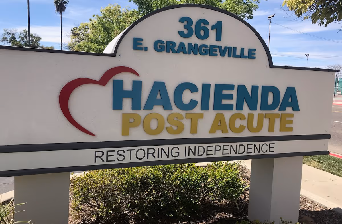 Outdoor sign for Hacienda Post Acute located at 361 E. Grangeville with a heart logo and the tagline 'Restoring Independence'. The sign is surrounded by greenery and a sidewalk is visible nearby.