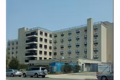 Exterior view of a multi-story beige building with multiple windows and balconies, several parked cars in front, and a clear sky above.