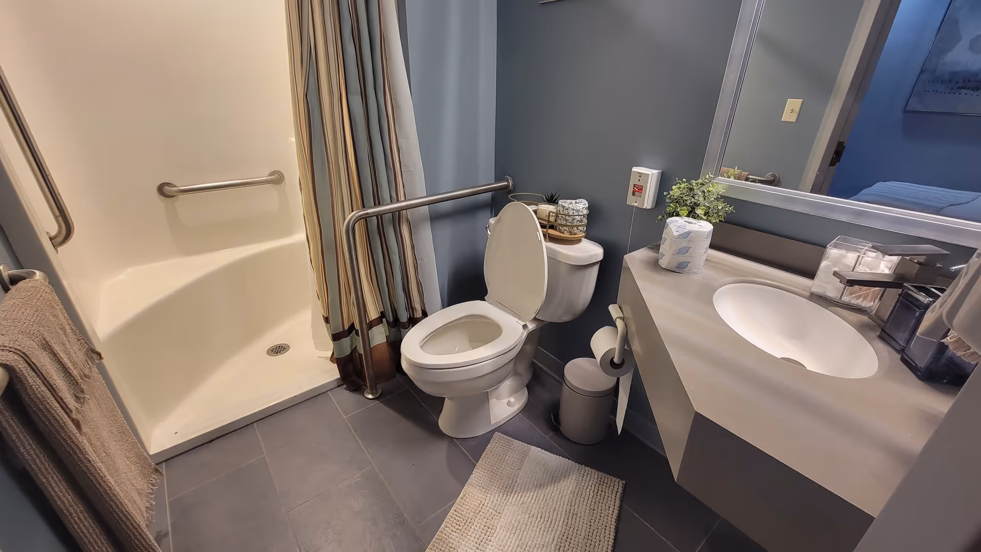 A bathroom with a toilet, a sink with a countertop, a large mirror, and a walk-in shower with a striped shower curtain. There are grab bars near the toilet and in the shower for accessibility. A towel hangs on a bar near the shower, and a small rug is placed on the tiled floor. The walls are painted blue, and there are some decorative items on the sink counter.