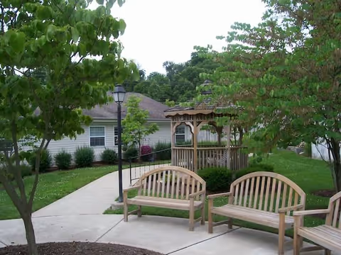 Outdoor garden area with wooden benches arranged around a paved walkway, a wooden gazebo in the background, green trees and shrubs, and a building with white siding visible behind the garden.