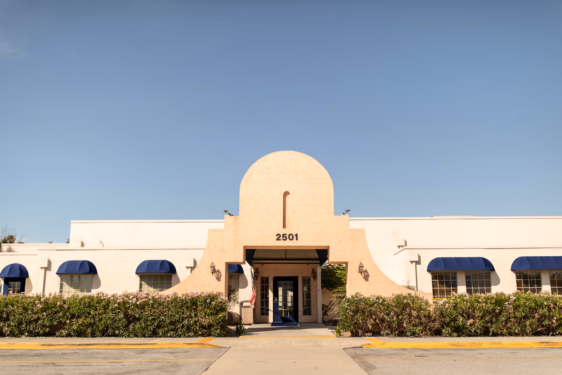 Front exterior view of a single-story building with a beige stucco facade and blue awnings over the windows. The building has an arched entrance with the number 2501 above the doorway, surrounded by green bushes and a clear blue sky in the background.