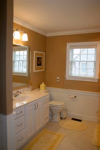 A bathroom with beige walls and white wainscoting, featuring a white vanity with a sink and mirror, a toilet, a window with white blinds, and yellow bath mats on the floor.