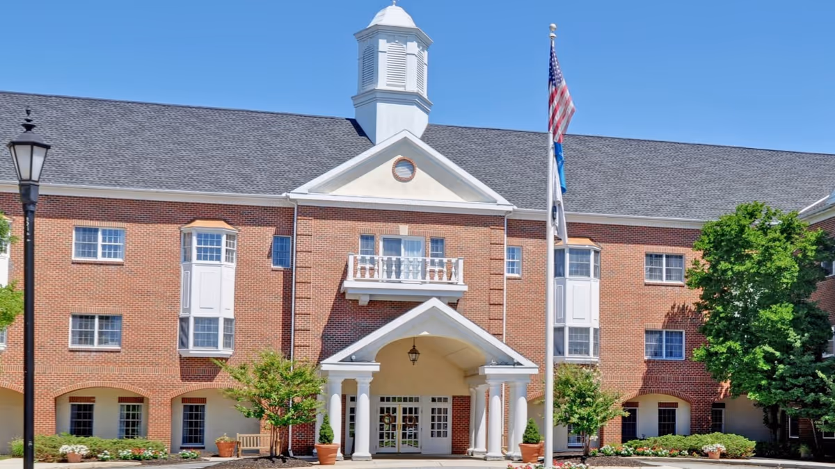 Front exterior view of a three-story senior living facility building with red brick walls, white columns at the entrance, a small balcony above the entrance, and a cupola on the roof. There are two flagpoles with flags in front of the building, along with some trees, shrubs, and a lamppost. The sky is clear and blue.