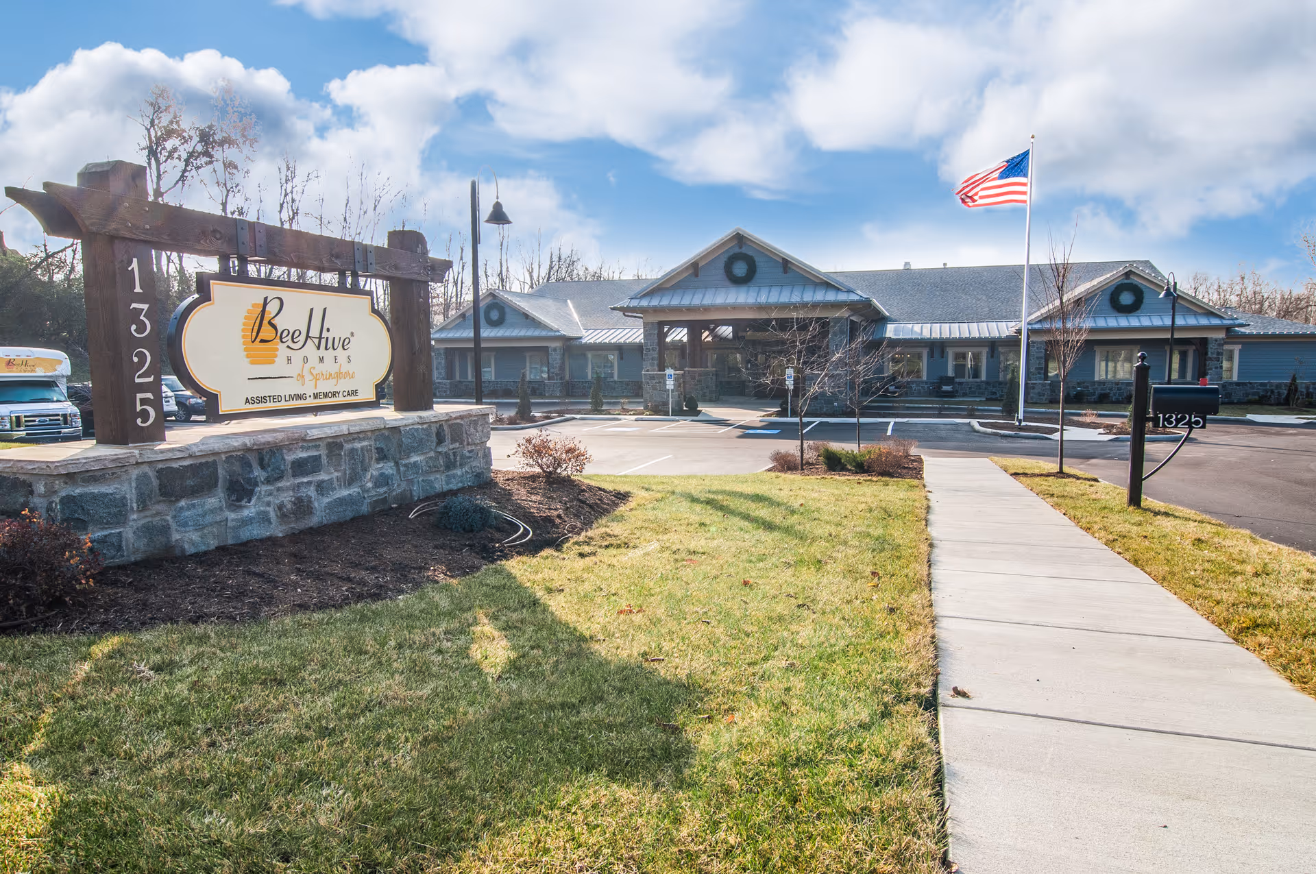 Exterior view of a senior living facility with a stone and wood sign reading 'BeeHive Homes of Springfield Assisted Living Memory Care' and the address number 1325. The building has a covered entrance, wreath decorations, an American flag on a flagpole, and a sidewalk leading to the entrance surrounded by grass and landscaping.