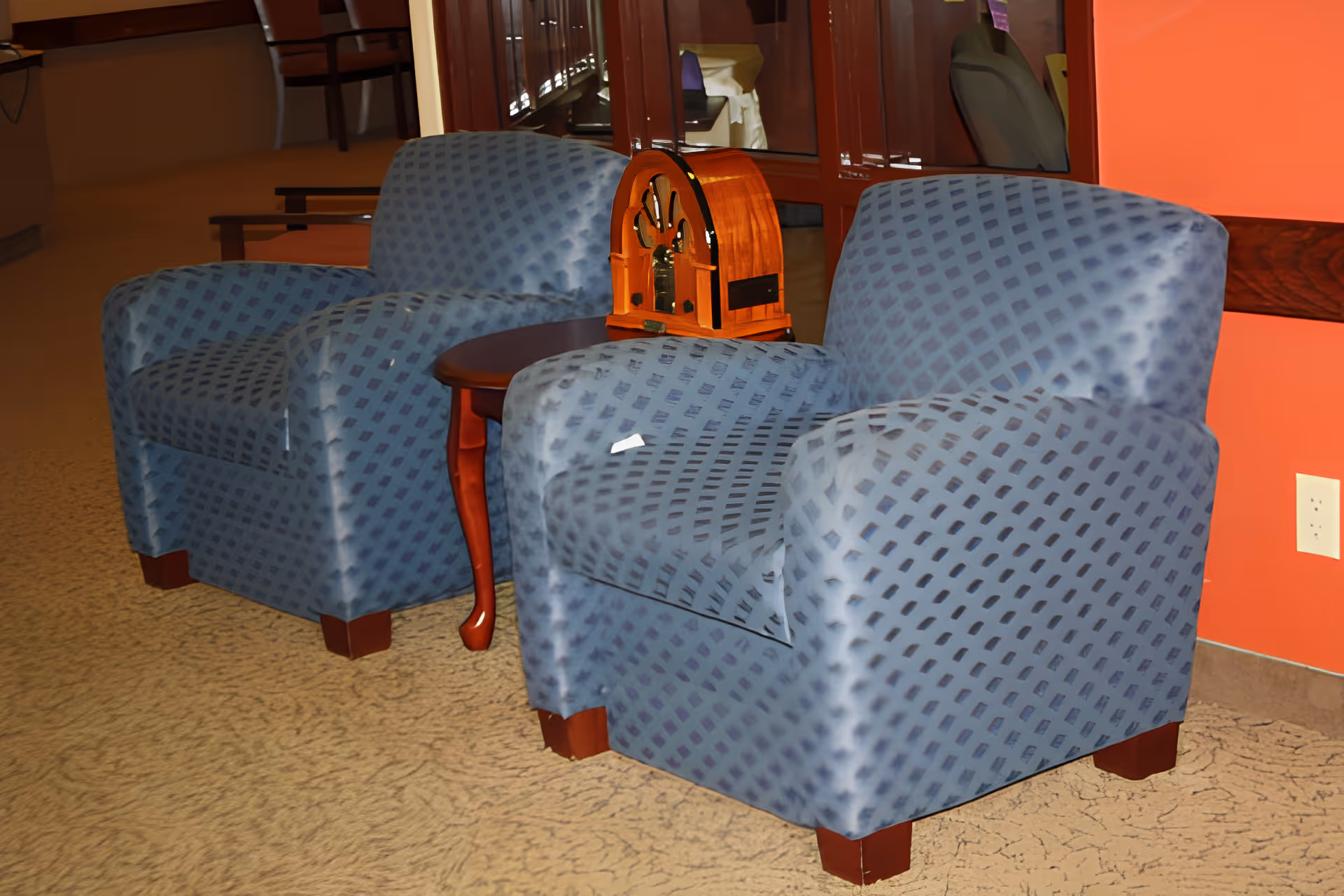 Two blue patterned armchairs with wooden legs are positioned around a small wooden side table. On the table sits a vintage-style wooden radio. The background includes a red wall with a white electrical outlet and a reflective glass surface showing part of the room.