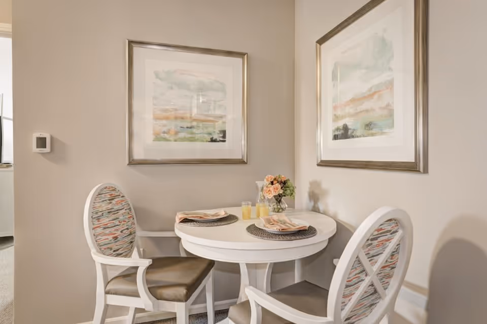 A small round white dining table set for two with placemats, napkins, two glasses of orange juice, and a vase of flowers. Two white chairs with patterned upholstery are positioned around the table. The walls are beige with two framed abstract paintings hanging above the table.