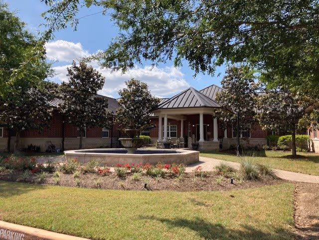 Exterior view of a single-story brick building with a metal roof, surrounded by trees and landscaping. In front of the building is a circular fountain with a stone border, and a walkway leading to the entrance under a covered porch with white columns. The sky is partly cloudy with blue patches visible.