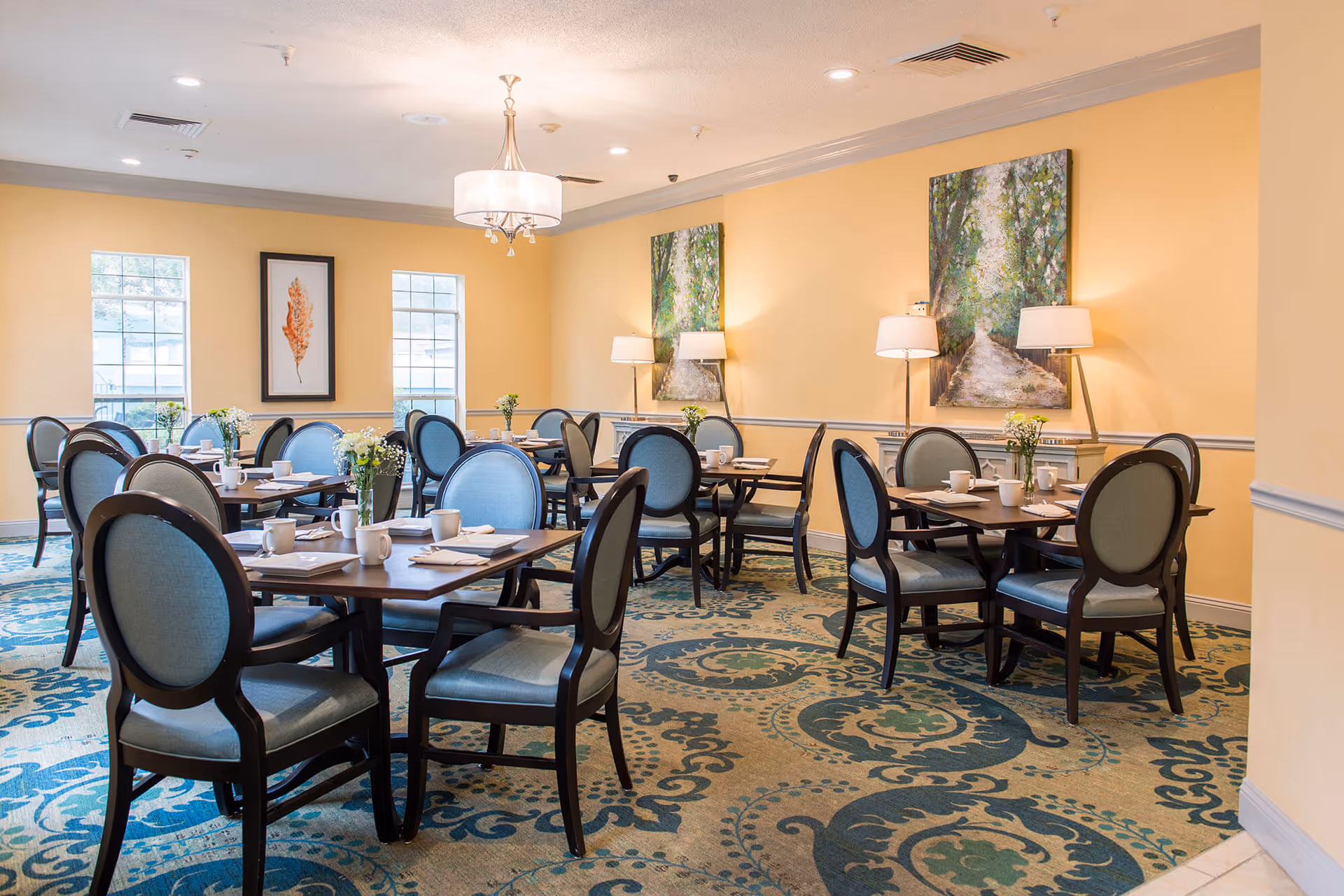 A bright dining room with several dark wood tables and blue upholstered chairs. Each table is set with white mugs, napkins, and small flower arrangements. The walls are painted yellow and decorated with framed artwork and lamps. The room has large windows allowing natural light to enter and a patterned carpet in shades of blue and green.