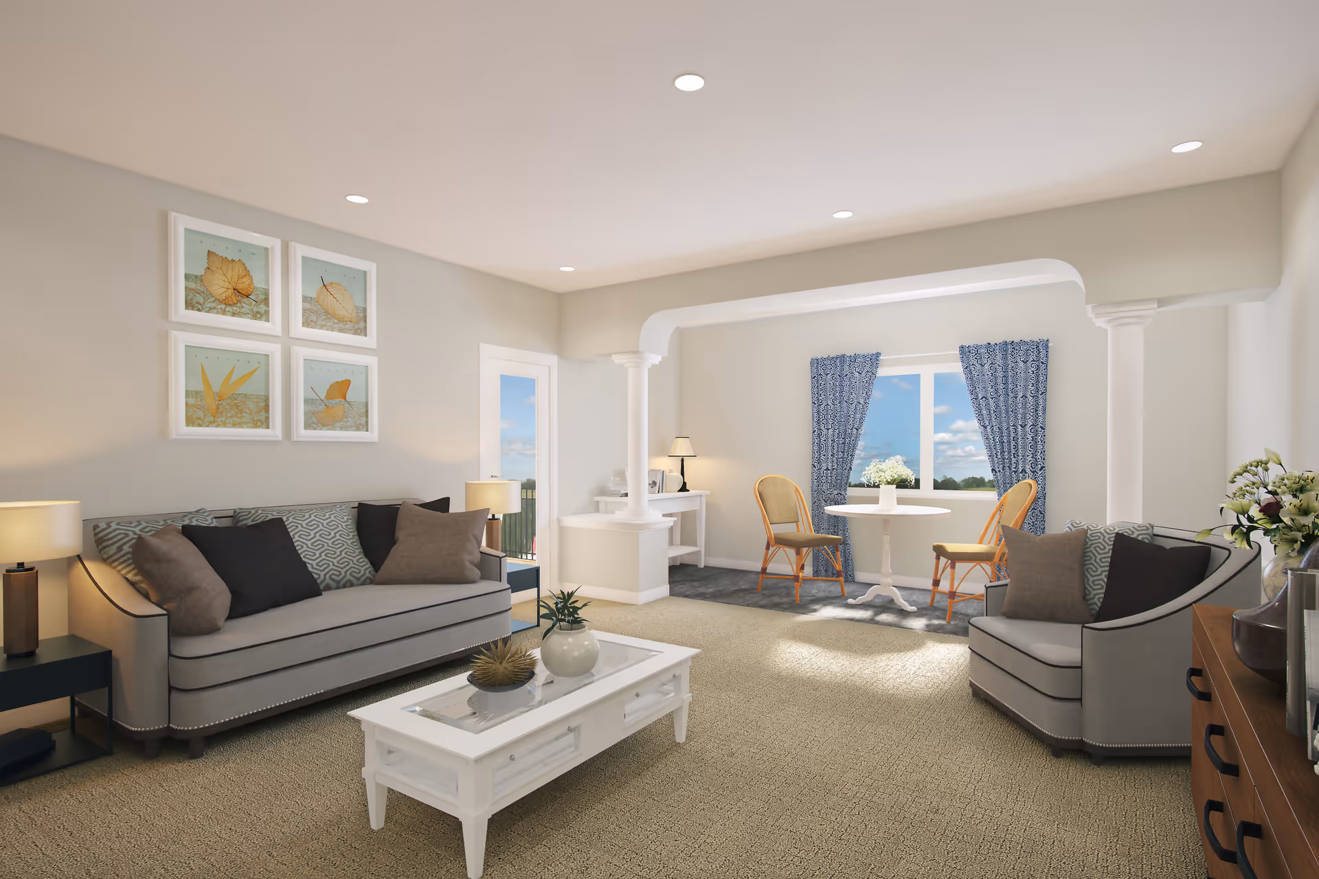 A bright and spacious living room with beige walls and carpet. The room features a gray sofa with multiple cushions, a matching armchair, a white coffee table with decorative items, and two side tables with lamps. There is a small round dining table with two wooden chairs near a window with blue patterned curtains. The room has recessed ceiling lights and framed artwork of leaves on the wall.