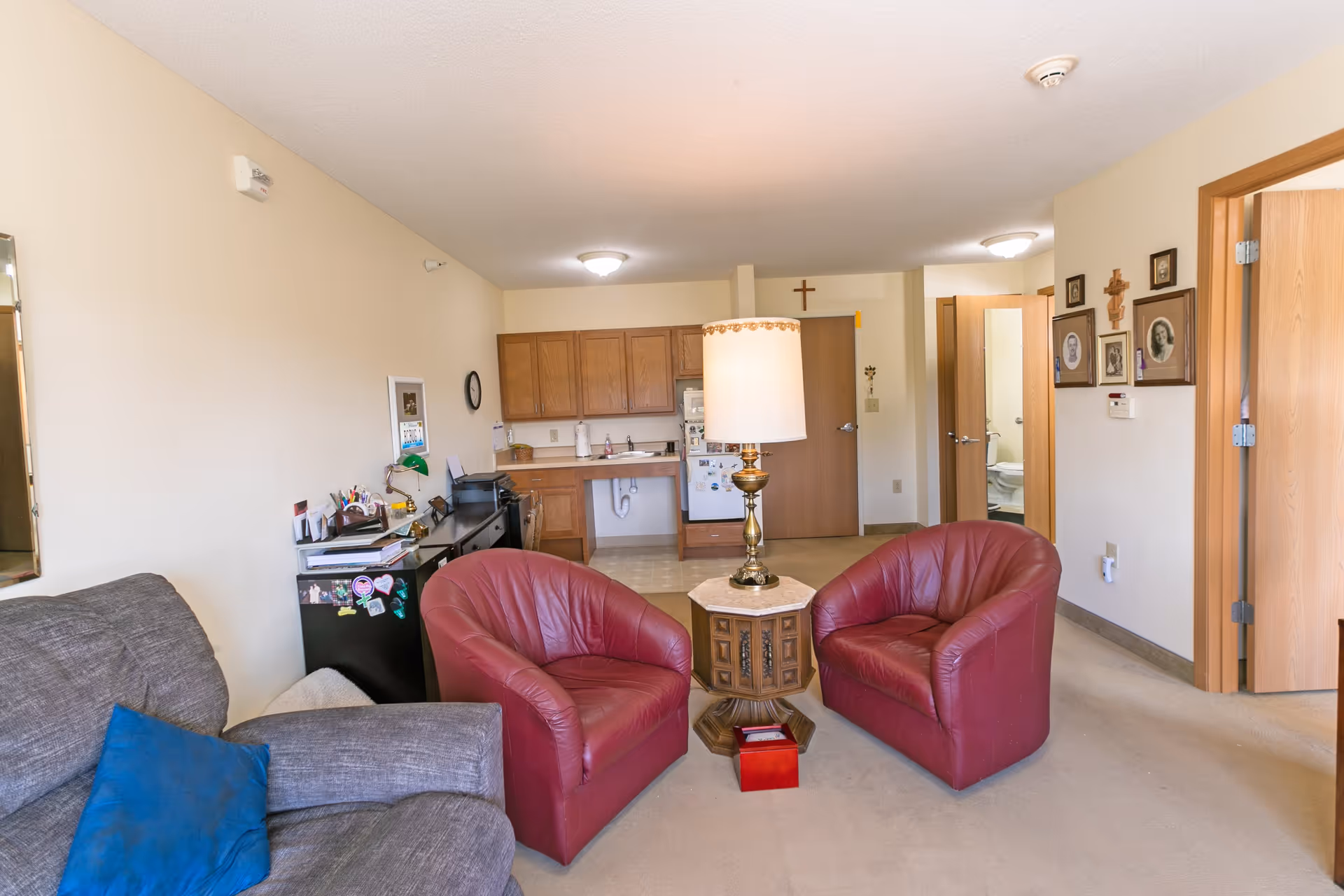 A cozy living area in a senior living facility featuring two red leather armchairs around a wooden side table with a lamp. To the left is a gray couch with a blue pillow. In the background, there is a small kitchenette with wooden cabinets, a mini fridge, and a sink. The walls are decorated with framed photos and crosses. An open door reveals a bathroom with a toilet.