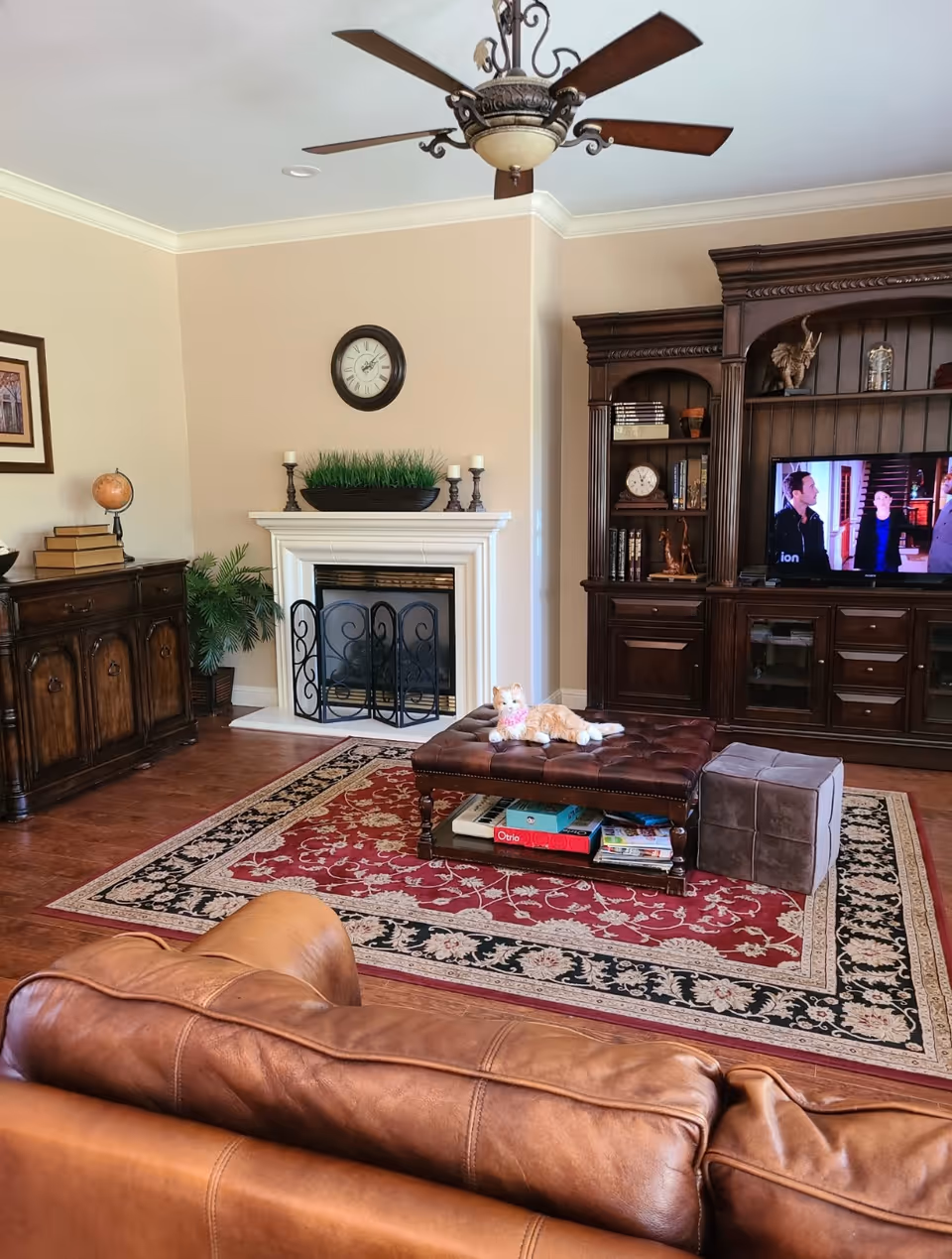 A cozy living room with a brown leather couch in the foreground, a red and beige patterned area rug, a tufted leather ottoman with a cat lying on it, a small gray cube ottoman, a fireplace with a decorative screen, a clock above the fireplace, a wooden cabinet with books and decorative items, a ceiling fan with wooden blades, and a TV showing a scene with people.