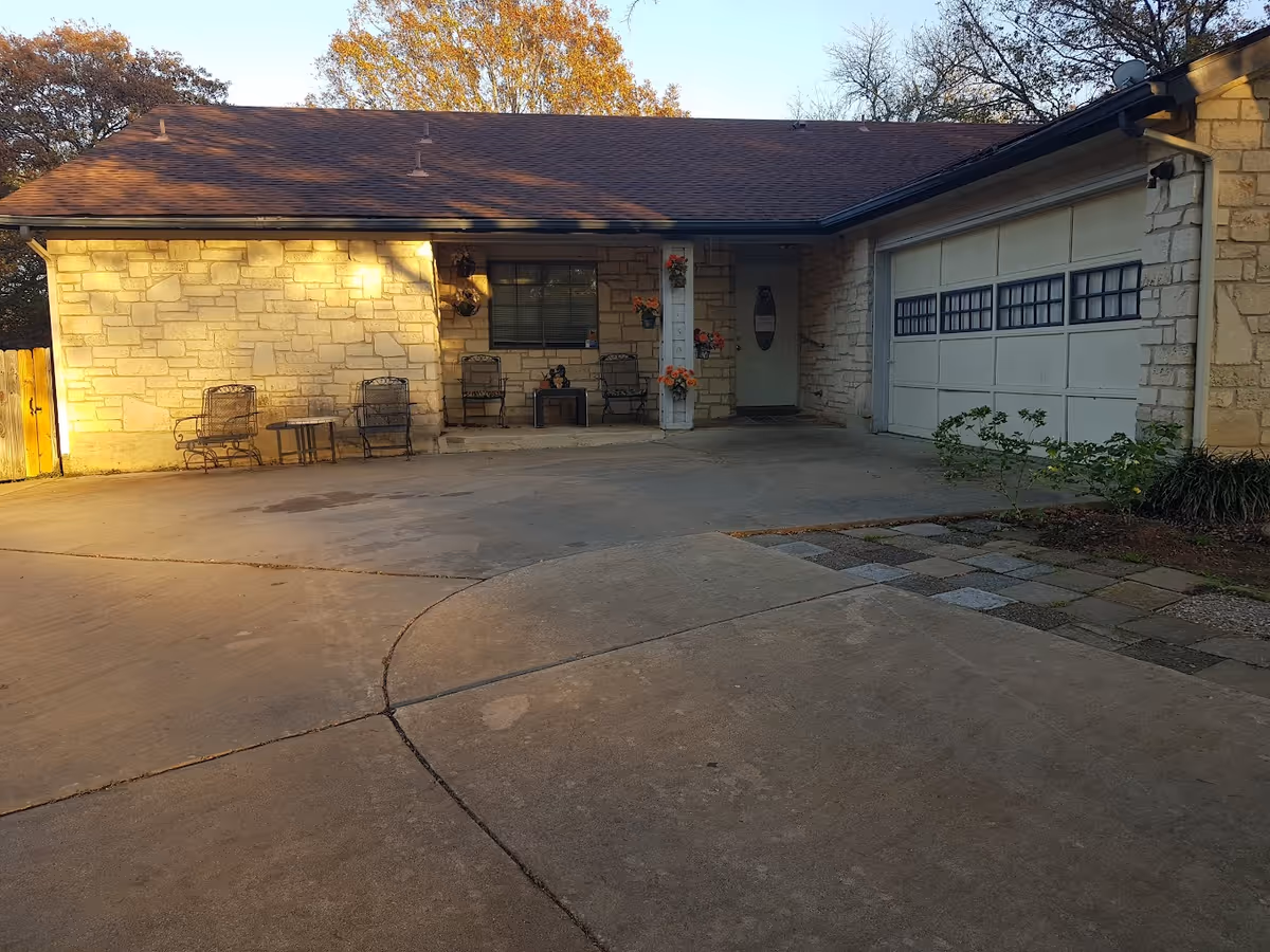 Exterior view of a single-story stone building with a brown shingled roof, a garage door on the right, and a small covered porch area with four metal chairs and a small table. There are flower pots with orange flowers hanging on the porch column and wall. The driveway and walkway are made of concrete and stone pavers, with some greenery on the right side.