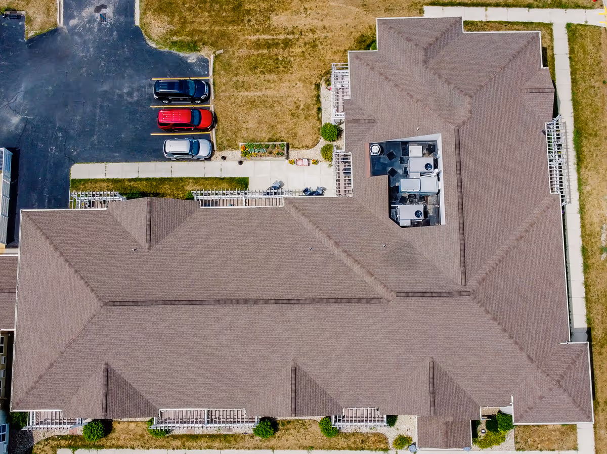 Aerial view of the roof and surrounding grounds of a senior living facility building with a parking lot containing three cars and adjacent sidewalks and grassy areas.