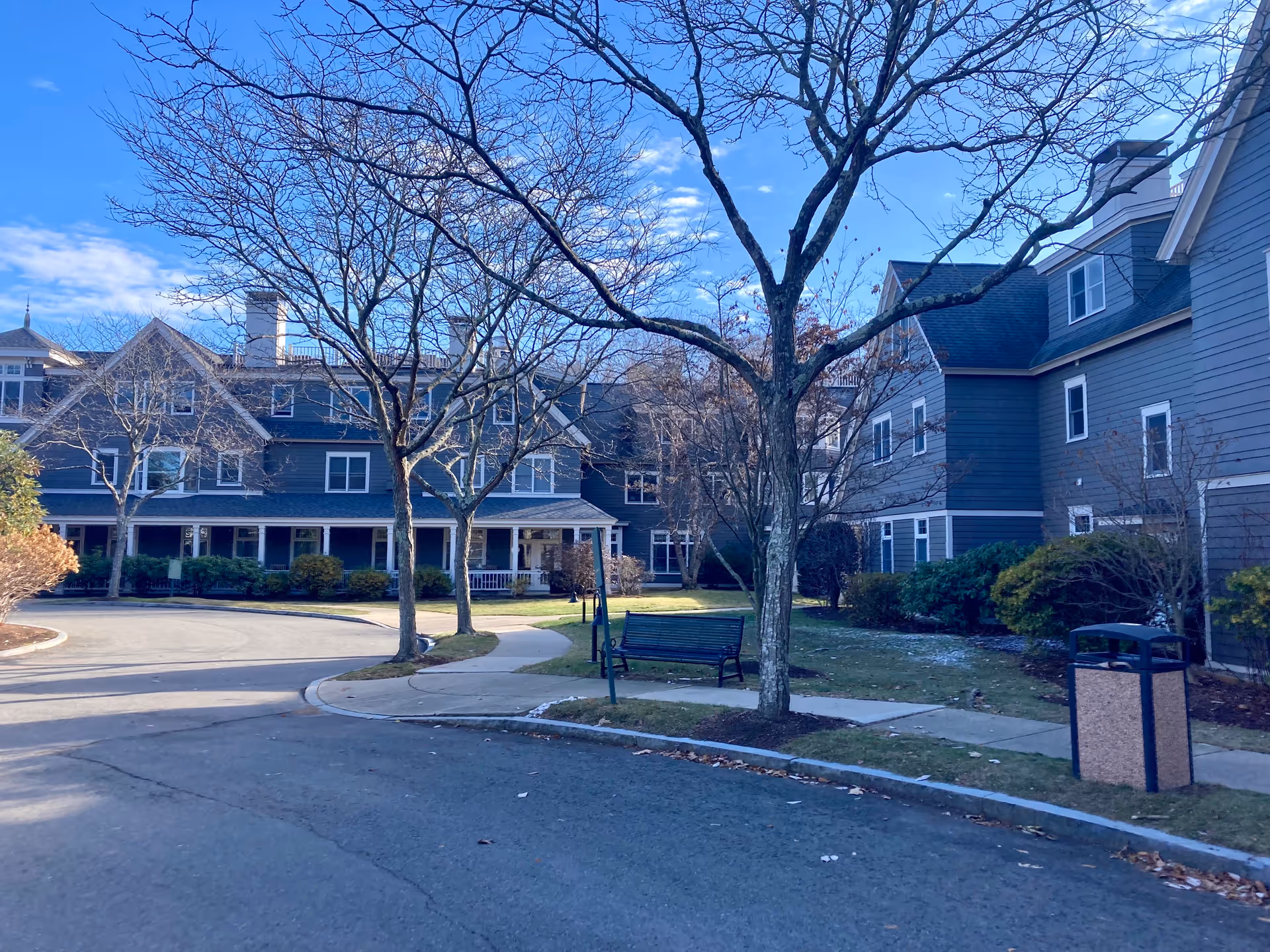 Exterior view of Goddard House Assisted Living facility showing a curved driveway, leafless trees, a bench, and a trash can. The building is multi-story with gray siding and white trim under a clear blue sky.