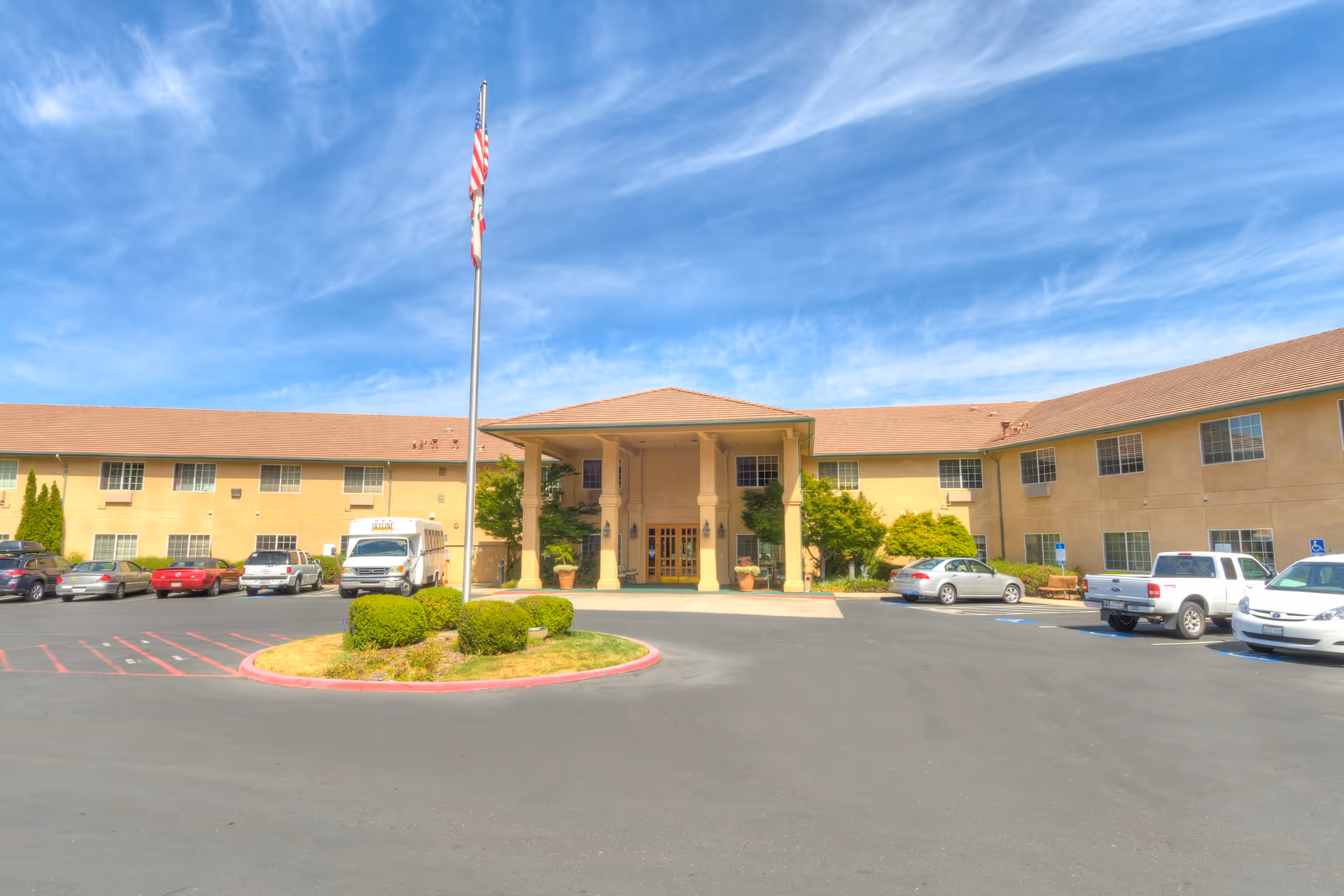 Exterior view of a two-story senior living facility building with a covered entrance, several parked cars, a flagpole with an American flag, and a clear blue sky with wispy clouds.