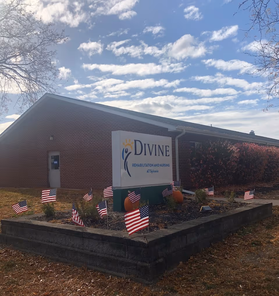 Exterior view of a brick building with a sign in front that reads 'Divine Rehabilitation and Nursing at Sylvania'. The sign is surrounded by small American flags and two pumpkins. The sky is partly cloudy with some trees visible on the sides.