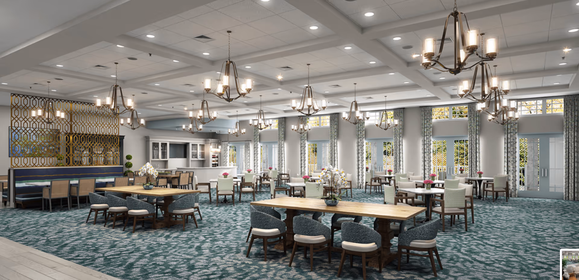 A spacious and well-lit dining room with multiple tables and chairs arranged neatly. The room features large windows with patterned curtains allowing natural light to enter. Elegant chandeliers hang from the ceiling, and decorative plants and flowers are placed on some tables. There is a partition with a geometric design separating part of the room, and a counter area is visible in the background.
