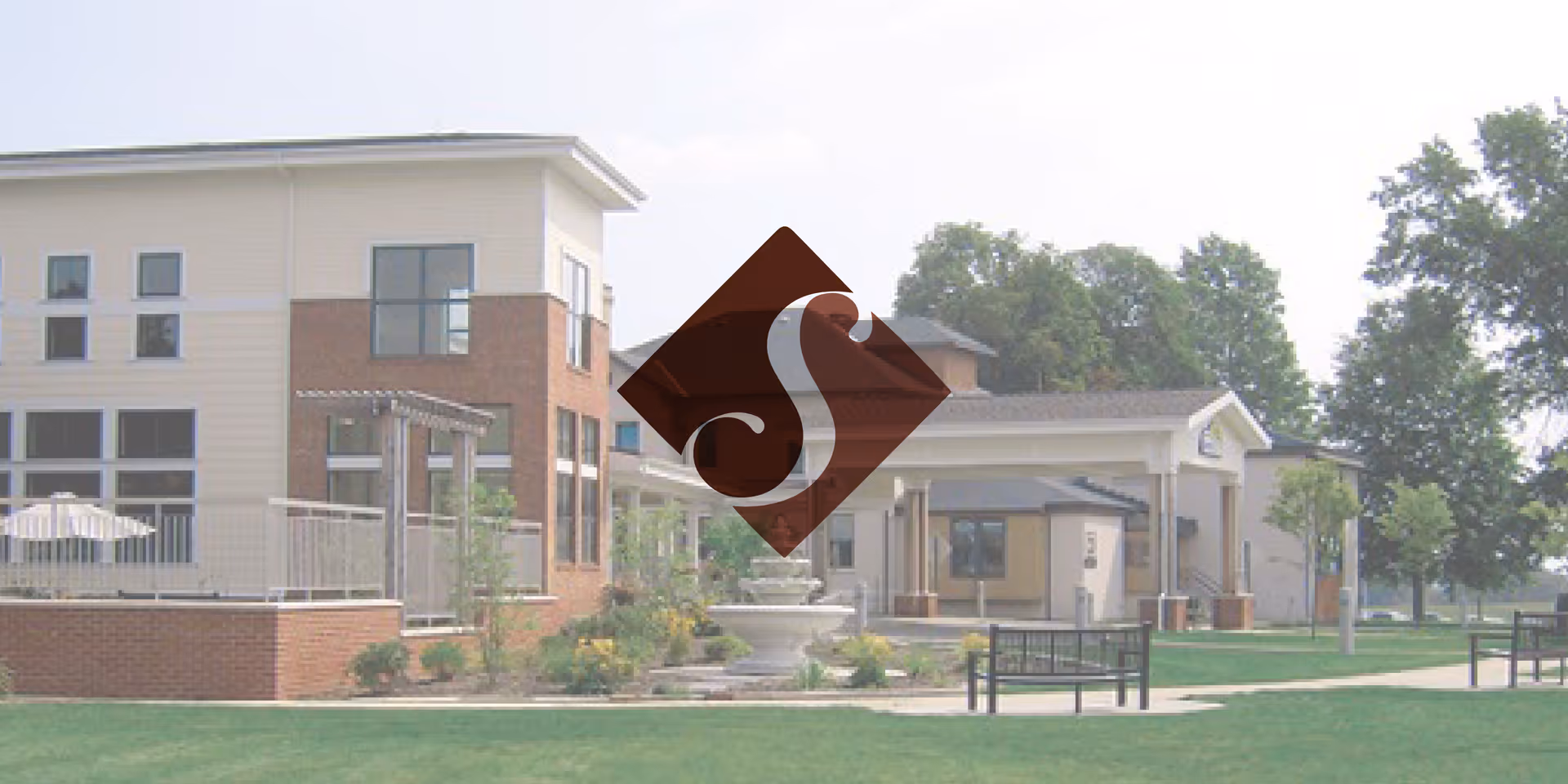 Exterior view of Sunnyview Nursing and Rehabilitation Center showing a modern building with large windows, a fountain in front, benches, and well-maintained green lawn with trees in the background.