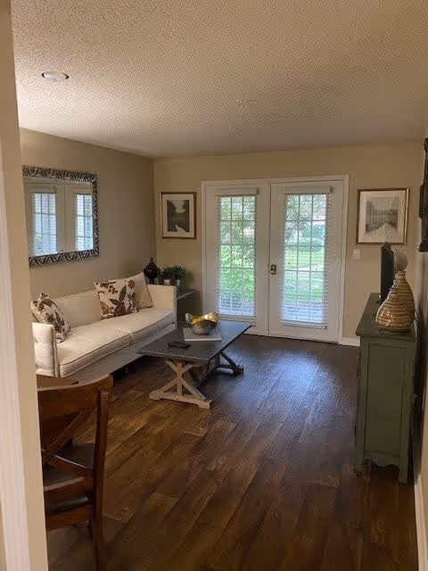 A cozy living room with a white couch adorned with patterned pillows, a wooden coffee table with decorative items, a large mirror on the wall, framed artwork, and double glass doors leading outside. The floor is wooden and there is a green cabinet with a vase on top.