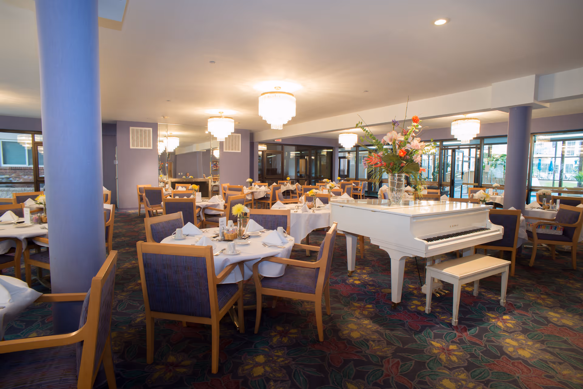 A spacious dining room with multiple round tables covered with white tablecloths, each set with napkins, cups, and small flower arrangements. A white grand piano with a large floral arrangement on top is positioned prominently in the room. The room features purple walls, patterned carpet with floral designs, wooden chairs with purple upholstery, and large windows letting in natural light. Several chandeliers hang from the ceiling providing warm lighting.