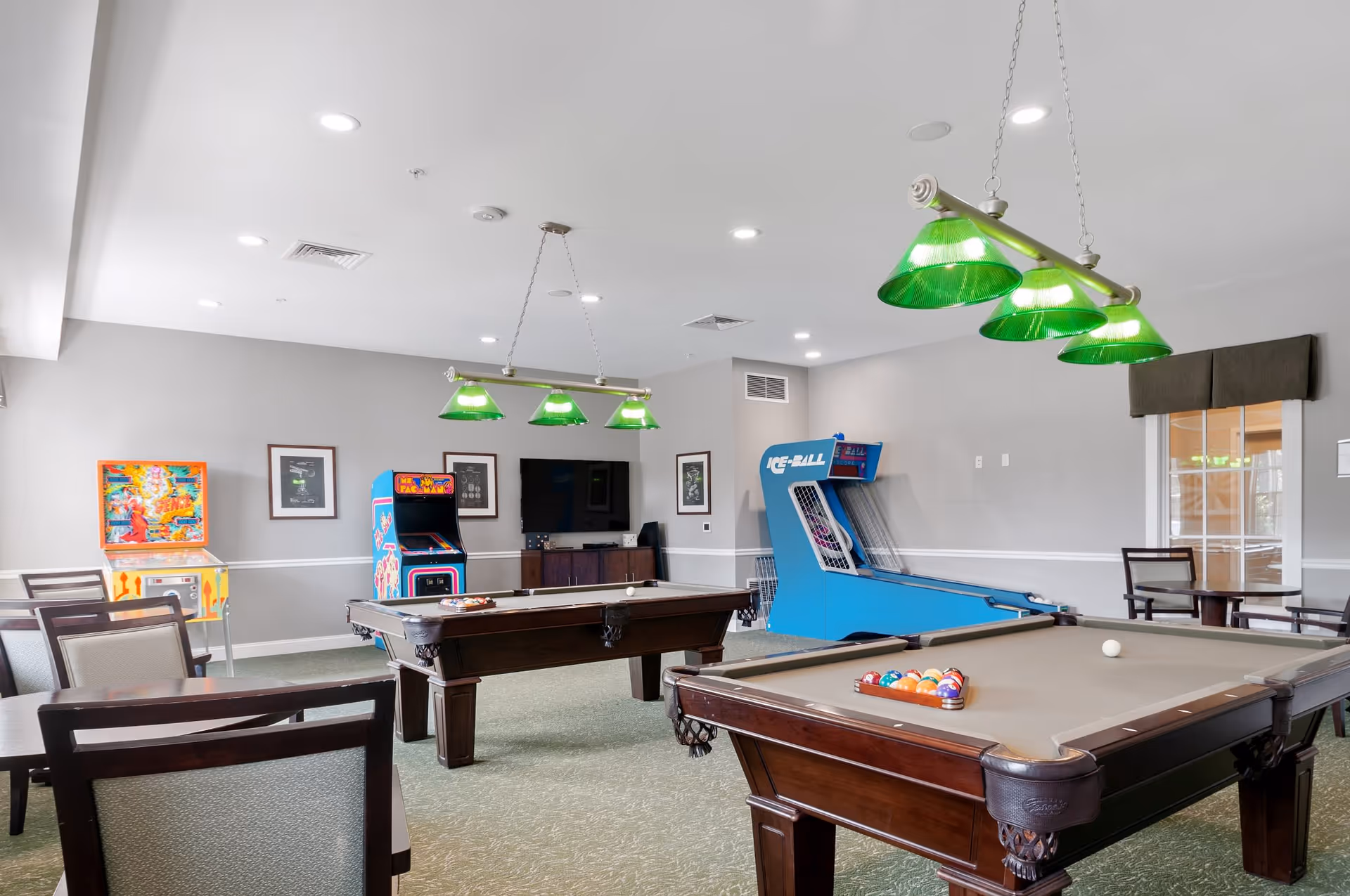 A recreational room with two pool tables, green hanging lights above them, arcade games including a Mr. Pac-Man machine and a skee-ball game, several chairs and tables, and a wall-mounted TV.