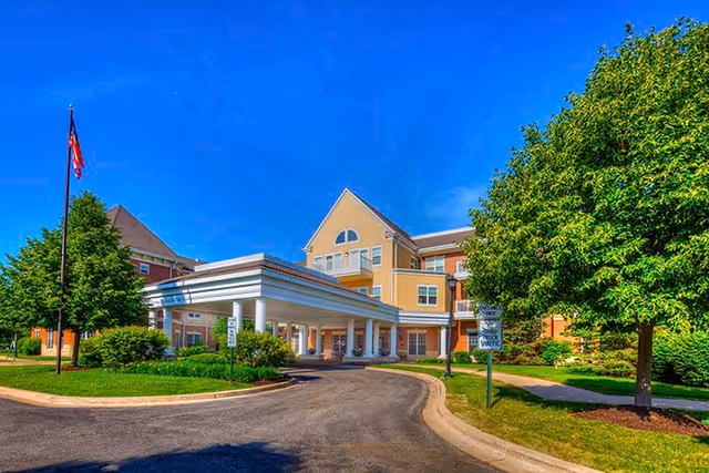 Exterior view of a senior living facility building with a covered entrance driveway, surrounded by green trees and landscaping under a clear blue sky.