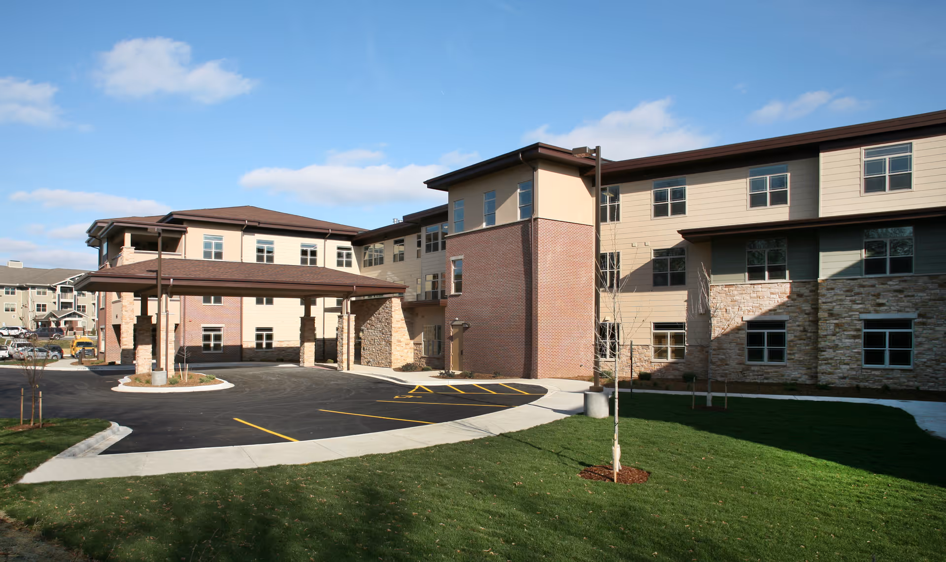 Exterior view of a modern senior living facility building with a covered entrance driveway, multiple windows, and a well-maintained lawn under a partly cloudy sky.