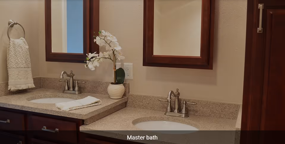 A master bathroom featuring two sinks with silver faucets set in a beige granite countertop. Above each sink is a wooden framed mirror. A white orchid plant in a white pot is placed between the sinks. A white hand towel hangs on a silver ring on the left wall, and another white towel is folded on the countertop.