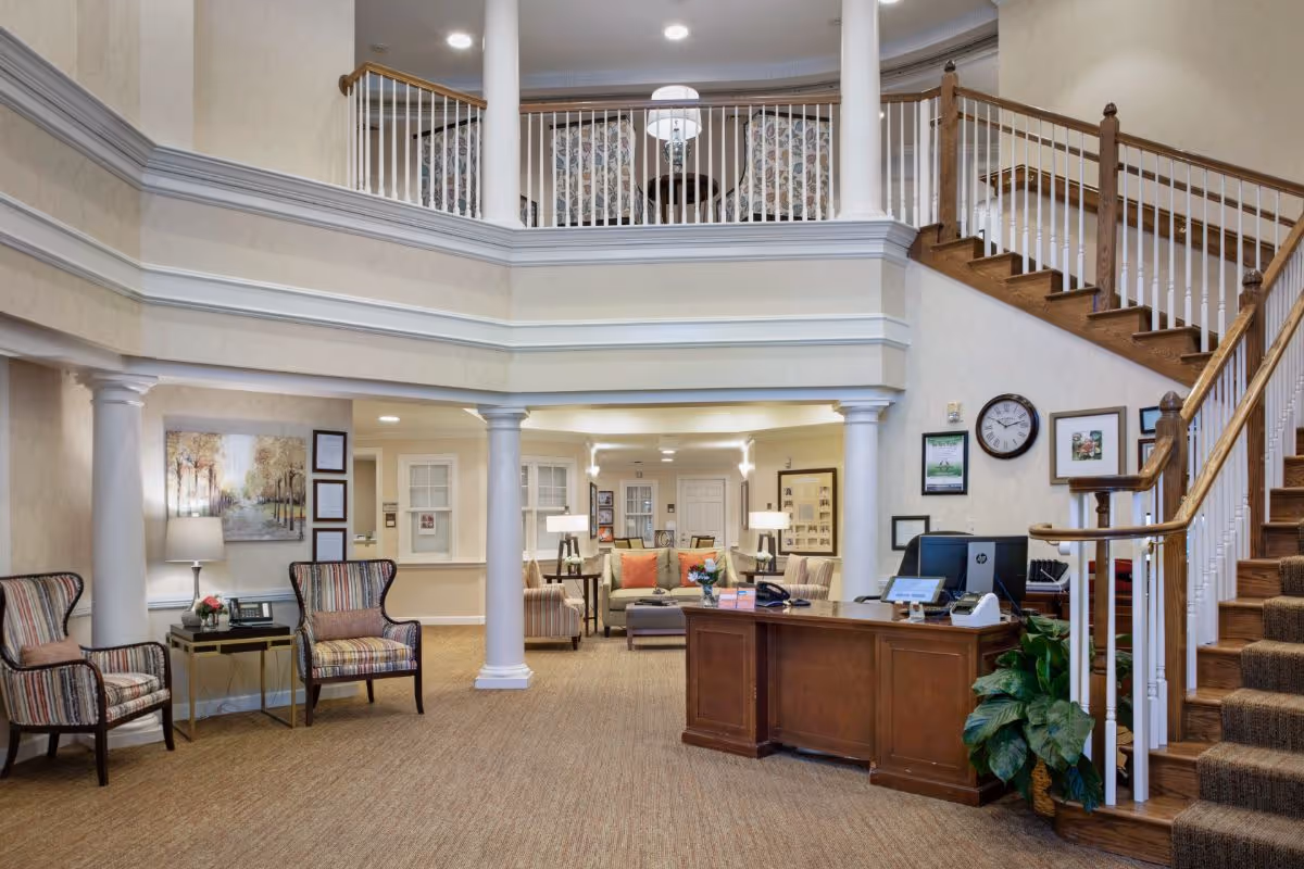 Interior view of a senior living facility lobby with a wooden reception desk, two white columns, a staircase with wooden handrails, and a seating area with striped chairs and a sofa. The walls are decorated with framed pictures and a clock, and there is a second-floor balcony with additional seating visible.