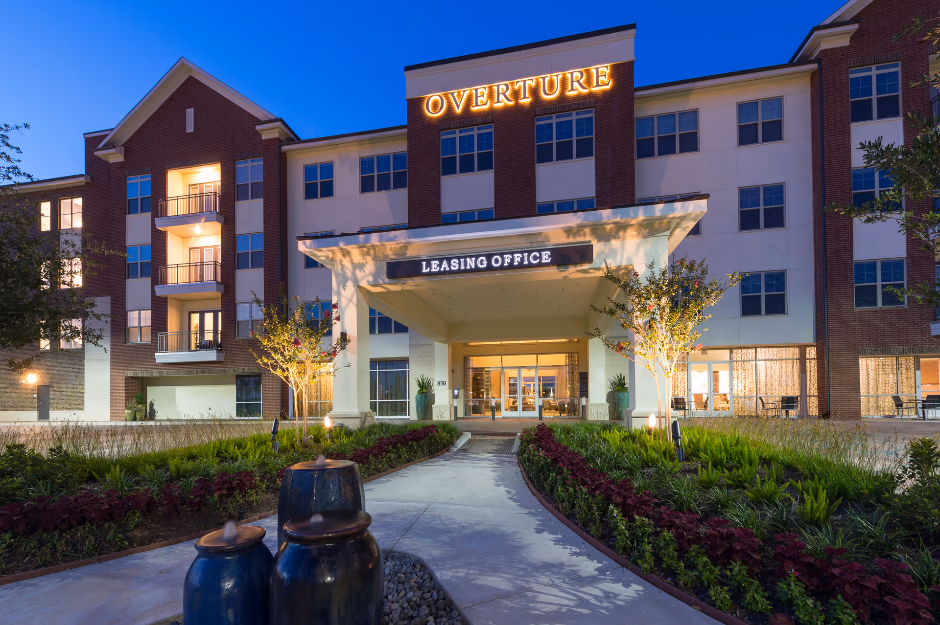 Exterior view of the Overture Sugar Land building at dusk with illuminated signage. The entrance features a covered walkway with a sign that reads 'Leasing Office.' There are landscaped gardens with plants and small trees along the pathway leading to the entrance. Three blue ceramic water fountains are visible in the foreground.