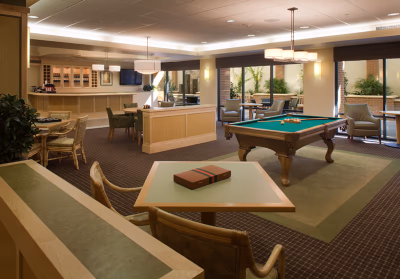A spacious common area in a senior living facility featuring a pool table, several tables and chairs, a bar area with stools, and large windows that let in natural light. The room is decorated with plants and has a warm, inviting atmosphere.
