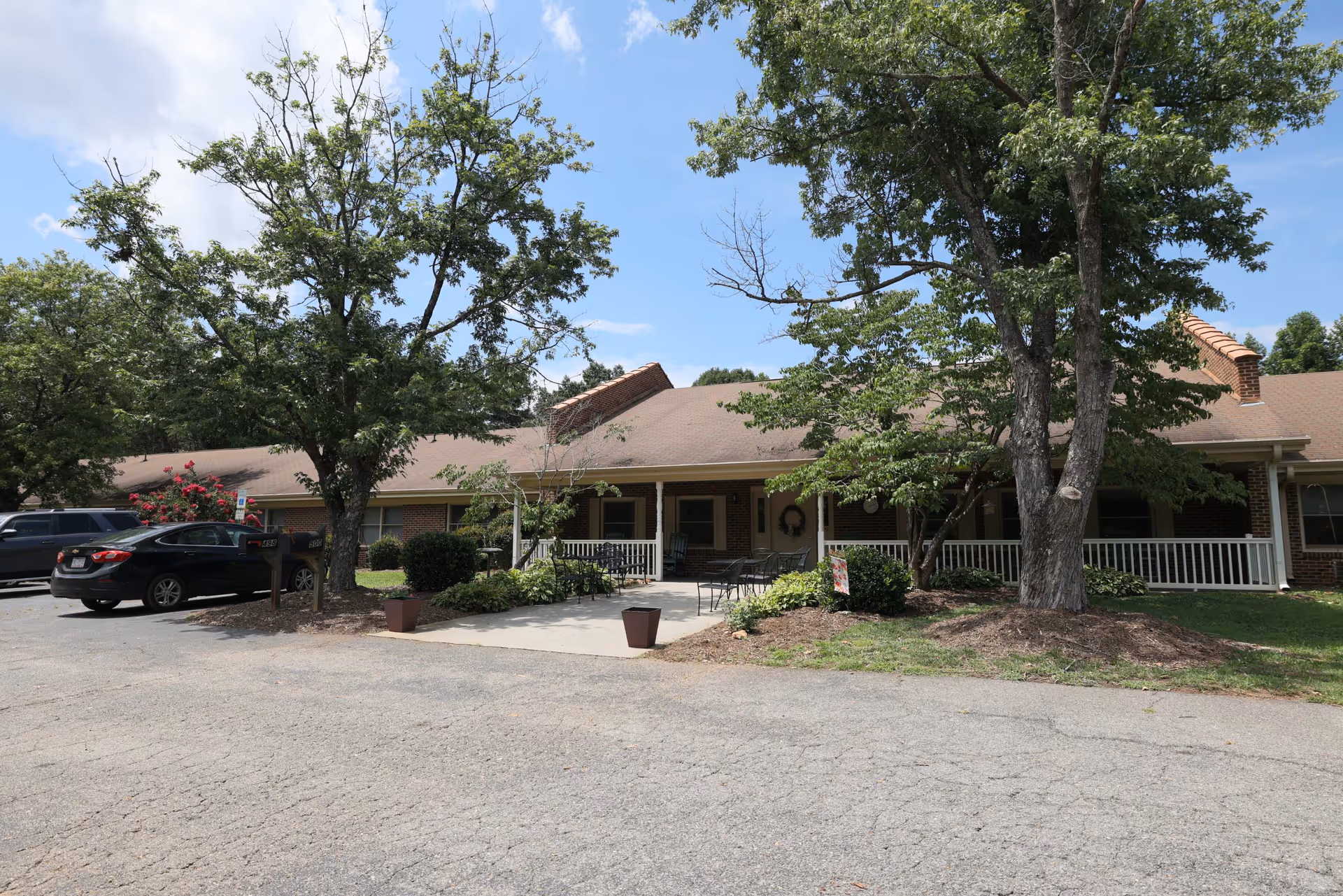 Exterior view of a single-story brick building with a covered porch and outdoor seating area, surrounded by trees and greenery under a partly cloudy sky. Several cars are parked in front of the building.
