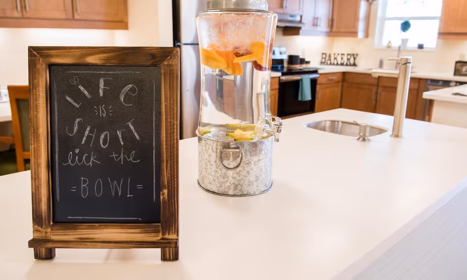 A kitchen countertop with a small chalkboard sign that reads 'Life is short lick the bowl' and a glass beverage dispenser filled with water and fruit slices. In the background, there are wooden cabinets, a stove, and a window with natural light.