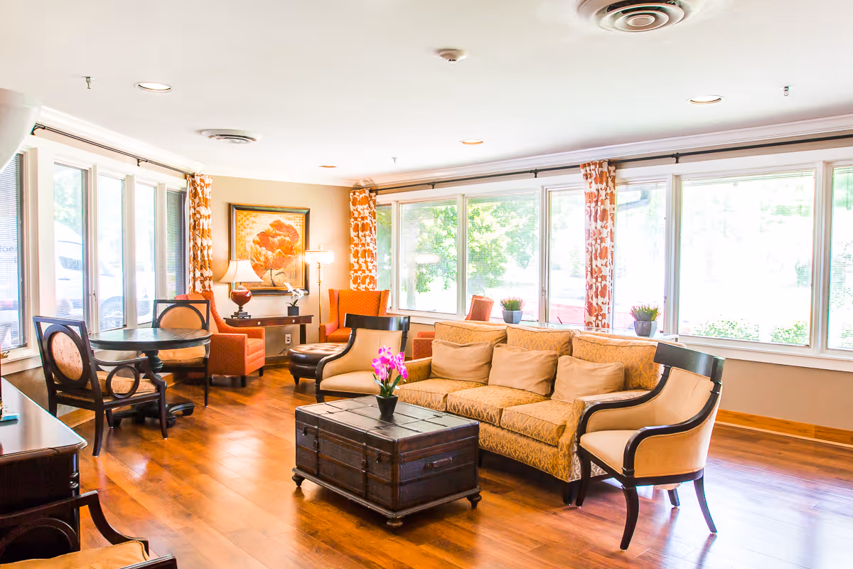 A bright and cozy living room area with large windows letting in natural light. The room features a patterned beige sofa with several cushions, two beige armchairs with dark wooden frames, a dark wooden coffee table with a small potted plant on top, and a round table with two chairs near the windows. The walls are decorated with orange patterned curtains and a framed floral painting. The floor is wooden, and there are two table lamps providing additional lighting.
