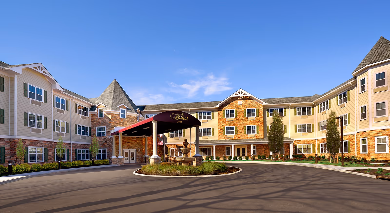 Front exterior view of The Bristal Assisted Living at Lake Grove building with a circular driveway, a covered entrance with a maroon canopy, and a fountain in the center of the driveway. The building has three stories with beige and brick facade, multiple windows, and a clear blue sky in the background.