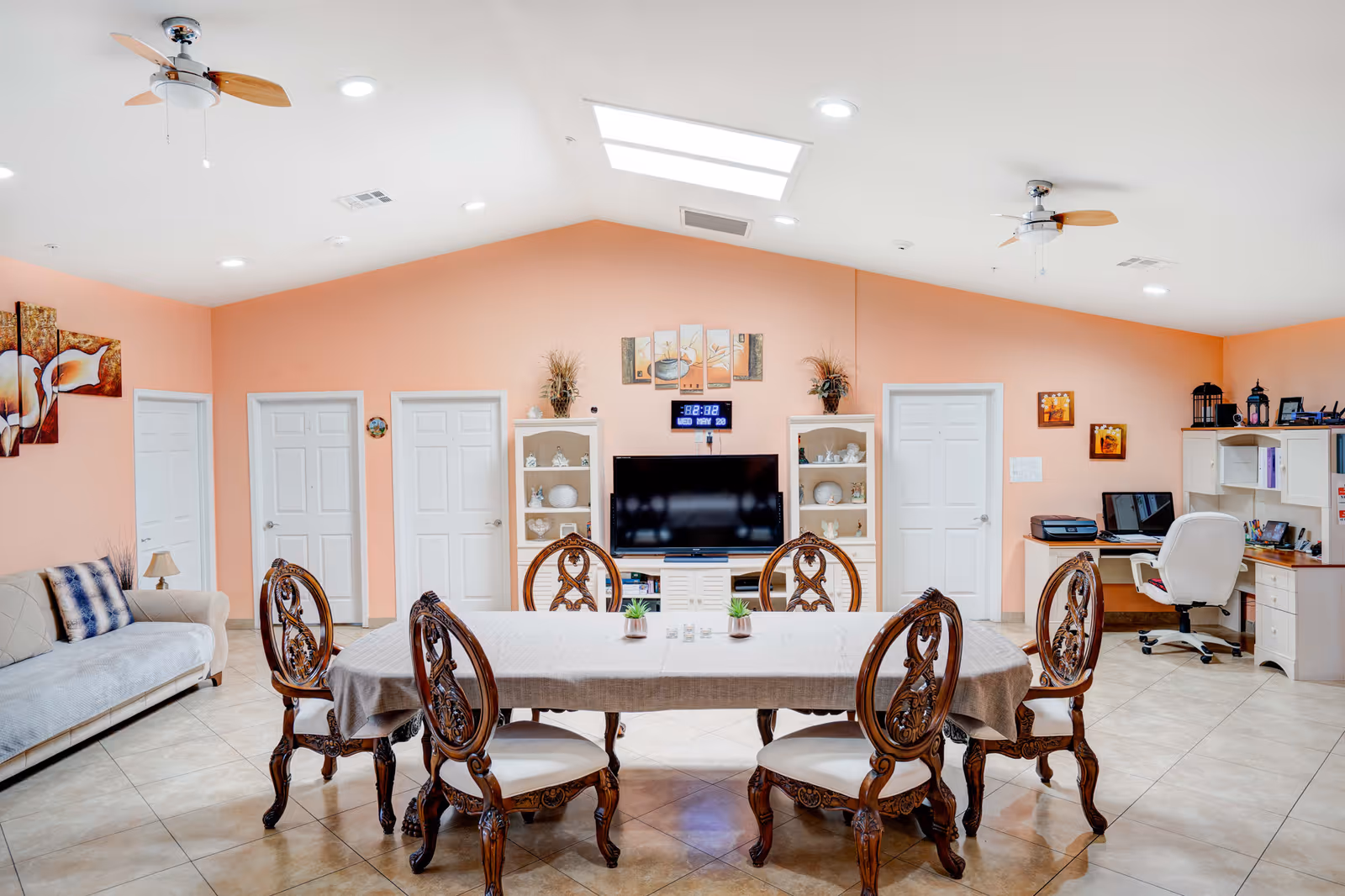 A spacious and well-lit common area with peach-colored walls, featuring a long dining table with six ornate wooden chairs covered with a beige tablecloth. There is a beige couch with a plaid pillow on the left side, a flat-screen TV mounted on a white cabinet with shelves displaying decorative items, and a work desk with a computer and office chair on the right side. The ceiling has two ceiling fans with wooden blades and a skylight.