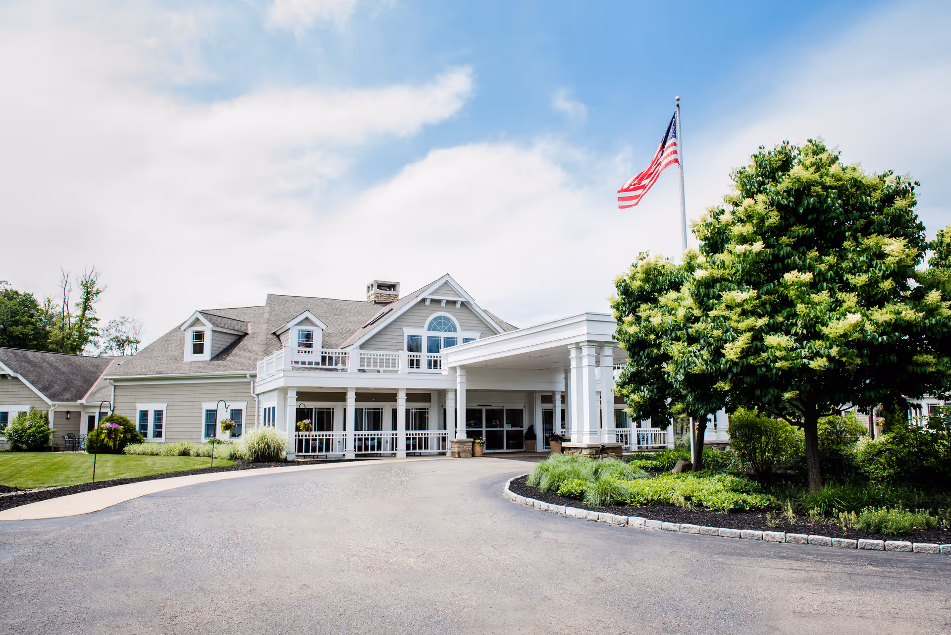 Front exterior view of a senior living facility building with a covered entrance, an American flag on a flagpole, well-maintained landscaping including green trees and shrubs, and a paved driveway leading to the entrance.