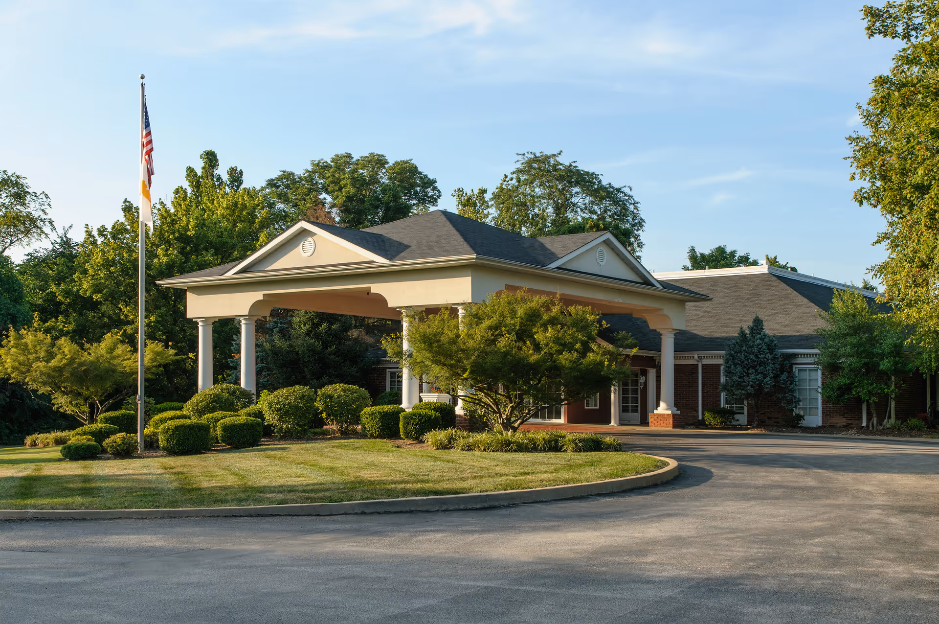 Exterior view of The Westchester House facility entrance with a covered driveway, surrounded by well-maintained bushes, trees, and a flagpole with an American flag, under a clear blue sky.
