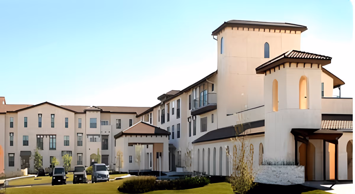 Exterior view of a large, multi-story senior living facility building with beige walls and brown roof tiles, featuring multiple windows and a covered entrance. There are several golf carts and a van parked on the driveway in front of the building, with some landscaping including small trees and bushes.