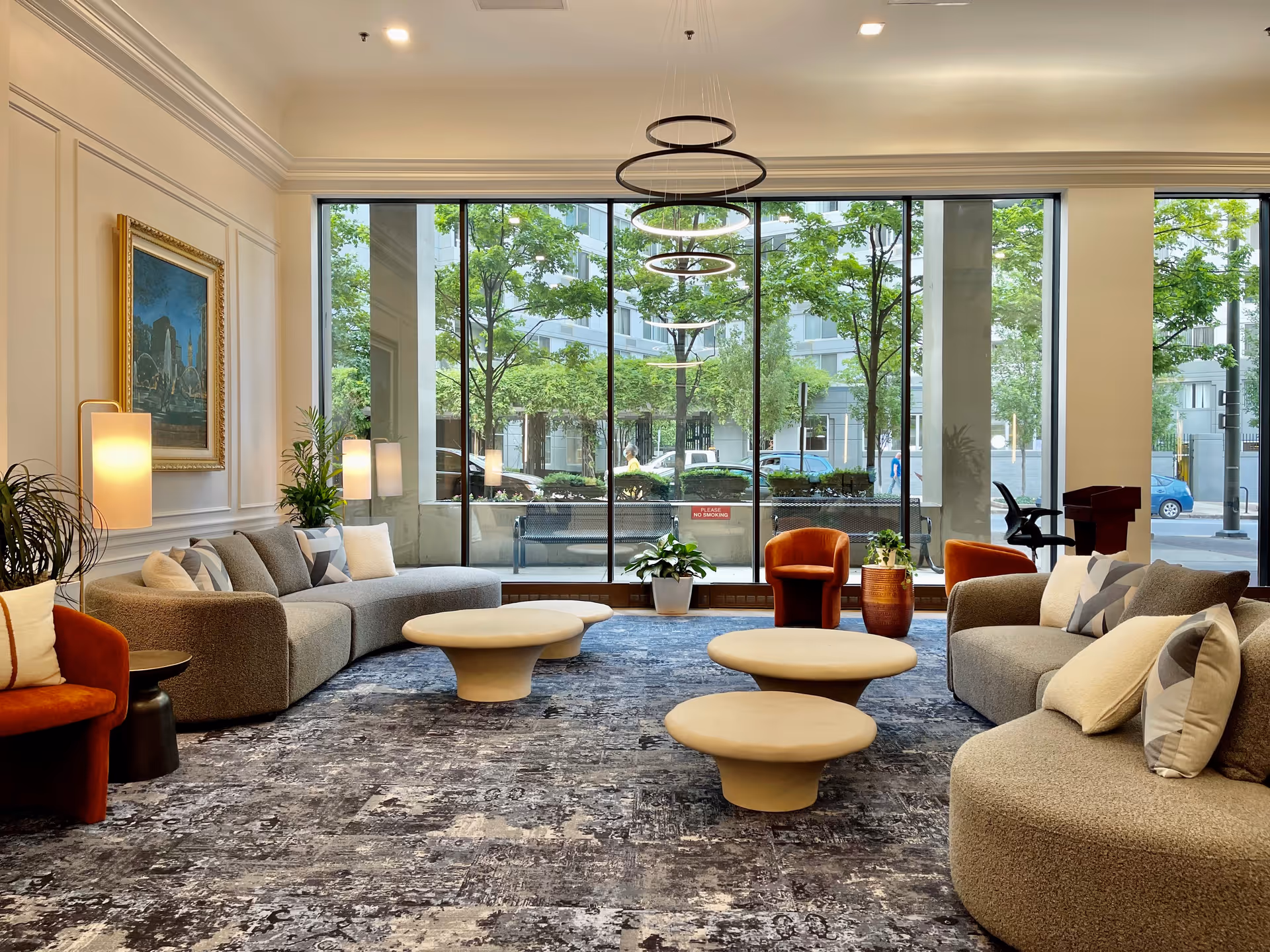 A modern and spacious living room with large floor-to-ceiling windows showing a view of trees and buildings outside. The room features curved gray sofas with multiple cushions, two orange armchairs, three round beige coffee tables, a patterned carpet, potted plants, a framed painting on the wall, and contemporary circular pendant lights hanging from the ceiling.