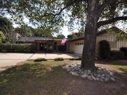 Front exterior view of a single-story building with a large tree surrounded by rocks in the foreground. The building has a garage, an American flag near the entrance, and is surrounded by greenery.