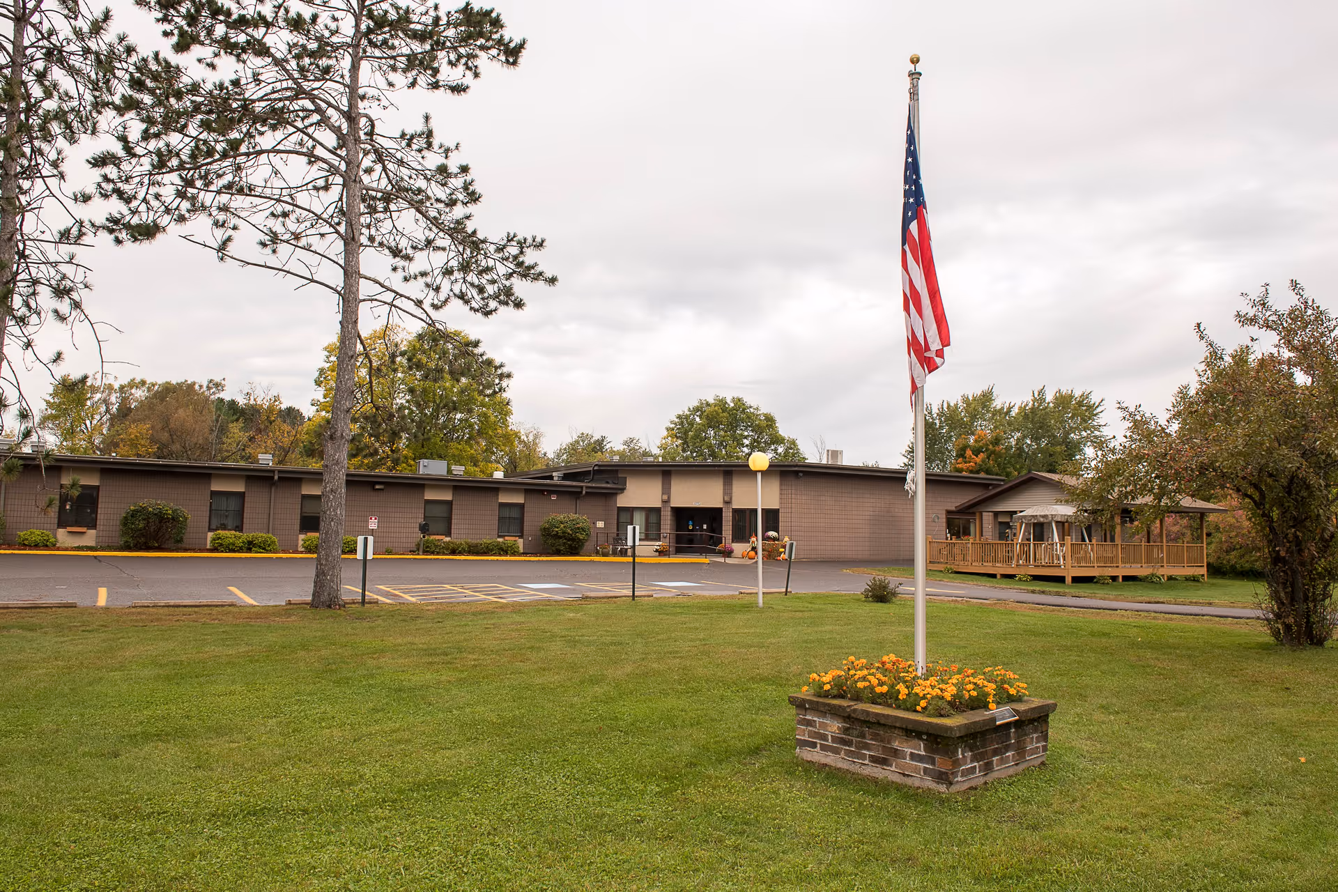 Exterior view of a single-story building with a parking lot in front, a flagpole with an American flag surrounded by flowers, and trees around the grassy area under a cloudy sky.