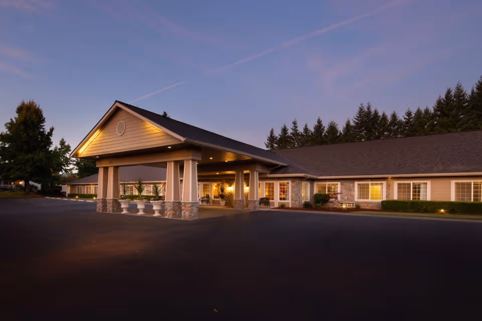 Exterior view of Brookstone Alzheimer's Special Care Center at dusk, showing a single-story building with a covered entrance, stone and siding facade, illuminated by warm lights, surrounded by trees and a paved parking area.