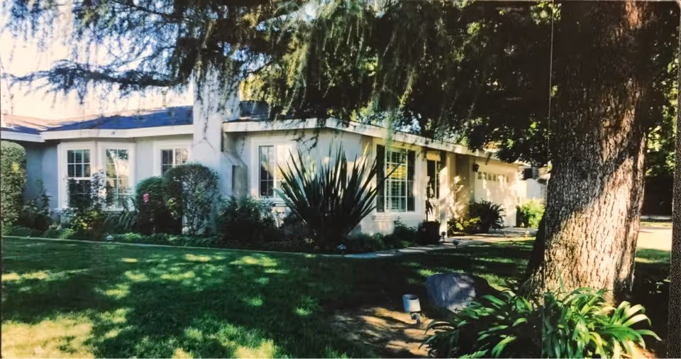 Exterior view of a single-story residential building surrounded by green grass, bushes, and large trees providing shade.