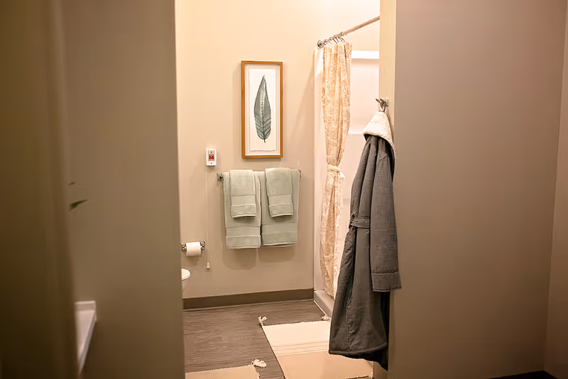View into a bathroom with beige walls, two green towels hanging on a towel rack, a framed leaf artwork above the towels, a shower with a beige patterned curtain, a gray robe hanging on a hook, and a beige rug on the floor.